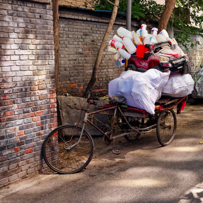A tricycle adapted with a large wooden frame extending from the front and sides, loaded with various discarded items including large white bags filled with waste, a children's red and black toy car, and numerous plastic bottles and containers stacked on top of the bags. The bicycle is positioned against a weathered brick wall on a paved surface, with a small tree and green foliage visible in the background. The scene depicts an informal method of rubbish collection or waste disposal, consistent with private waste handling and alternative rubbish removal methods, as part of a local waste management effort by Rubbish Removal Camberwell. The lighting is natural, likely in daylight, illuminating the textured brickwork, the metal and wood of the makeshift carrier, and the assorted waste materials, emphasizing the essential role of domestic waste management in urban environments.