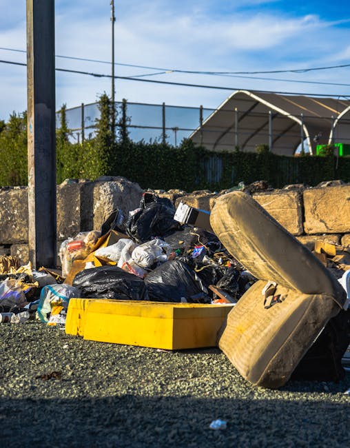 A close-up view of a pile of mixed waste and rubbish discarded on a gravel surface, including black plastic bin bags, a strewn yellow plastic container, a discarded old car tire leaning against the bags, and various smaller debris. Behind the waste, there is a low stone wall constructed of rough-hewn stones, and further in the background, a metal fence with greenery and trees visible beyond, along with a nearby sports facility or shelter with a curved roof structure. The scene is set outdoors under a bright, partly cloudy sky, and the image portrays an area awaiting waste collection or removal, typical of private rubbish disposal services such as those offered by Rubbish Removal Camberwell, focused on efficient on-site clearance of bulky refuse.