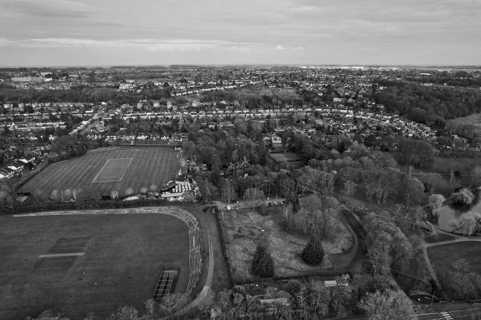 Aerial black-and-white photograph capturing a large urban park area surrounded by residential housing and trees, with a sports field featuring a running track, football pitch, and adjacent sports facilities in the foreground. The park includes pathways, landscaped sections with bushes and smaller trees, and a small pond with a curved shoreline on the right side of the image. The background reveals a densely built suburban area with rows of houses, streets, and a distant horizon, under a partly cloudy sky. The scene presents a quiet and expansive public space within a residential neighbourhood, indicative of a typical urban park, which could be relevant to private waste removal or on-site clearance services provided by companies like Rubbish Removal Camberwell operating near Myatts Field Park.