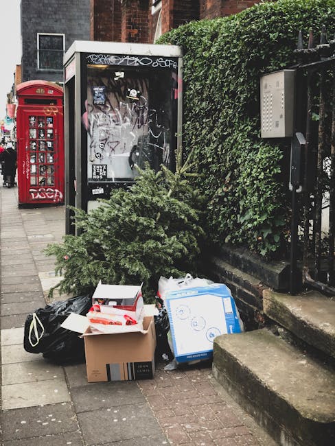 A small pile of rubbish on the pavement outside a red letterbox and a glass shelter, including a green artificial Christmas tree, packaged cardboard boxes, and black and white bags. The scene is set on a grey brick sidewalk next to a building with a hedge and black metal fence, with a payphone box and residential windows in the background. The rubbish appears to have been left temporarily and is in front of a white utility box attached to the fence. The overall setting suggests a typical urban streetscape where private rubbish removal services, such as those offered by Rubbish Removal Camberwell, might be used for alternative waste handling or on-site clearance.